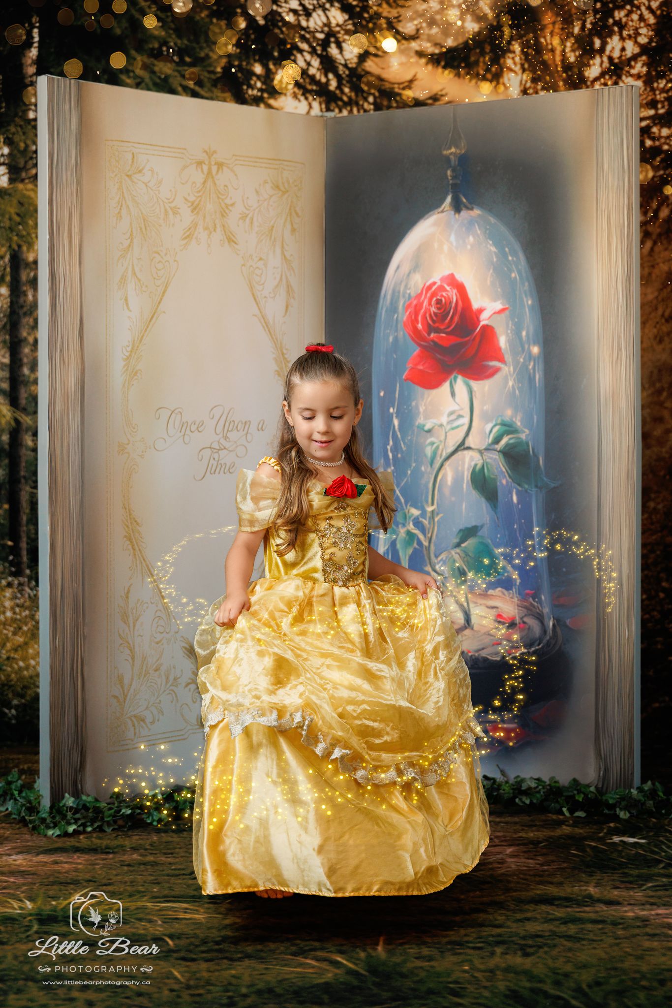 A young girl in a golden Belle princess gown poses before a Beauty and the Beast storybook backdrop with enchanted rose.