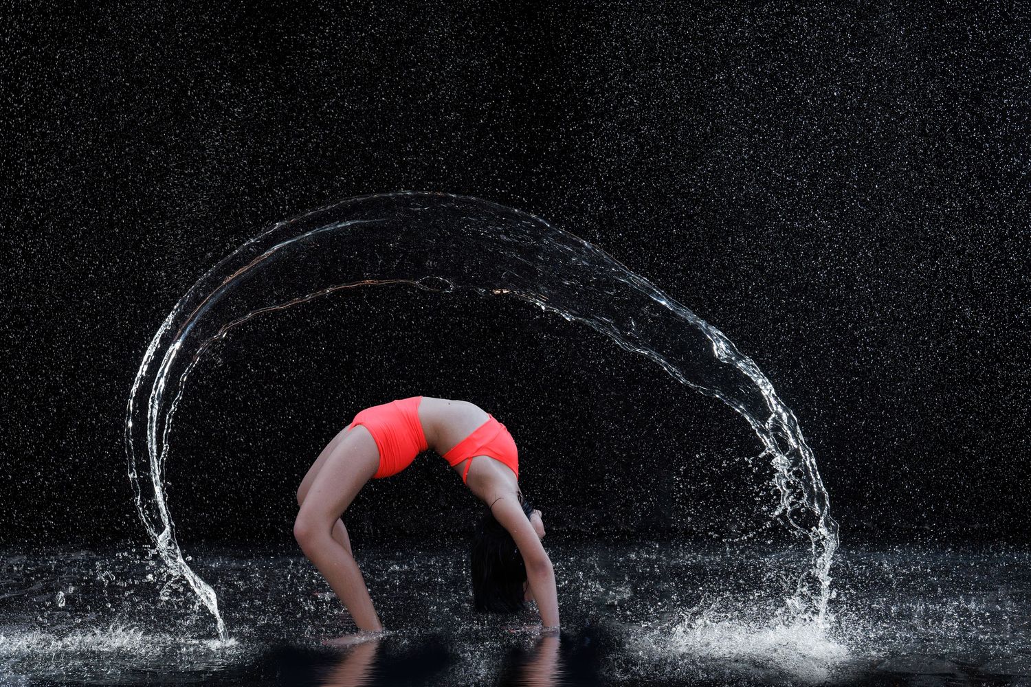 Gymnast in red bikini performs a backbend with water arcing gracefully around her body against a dark background.