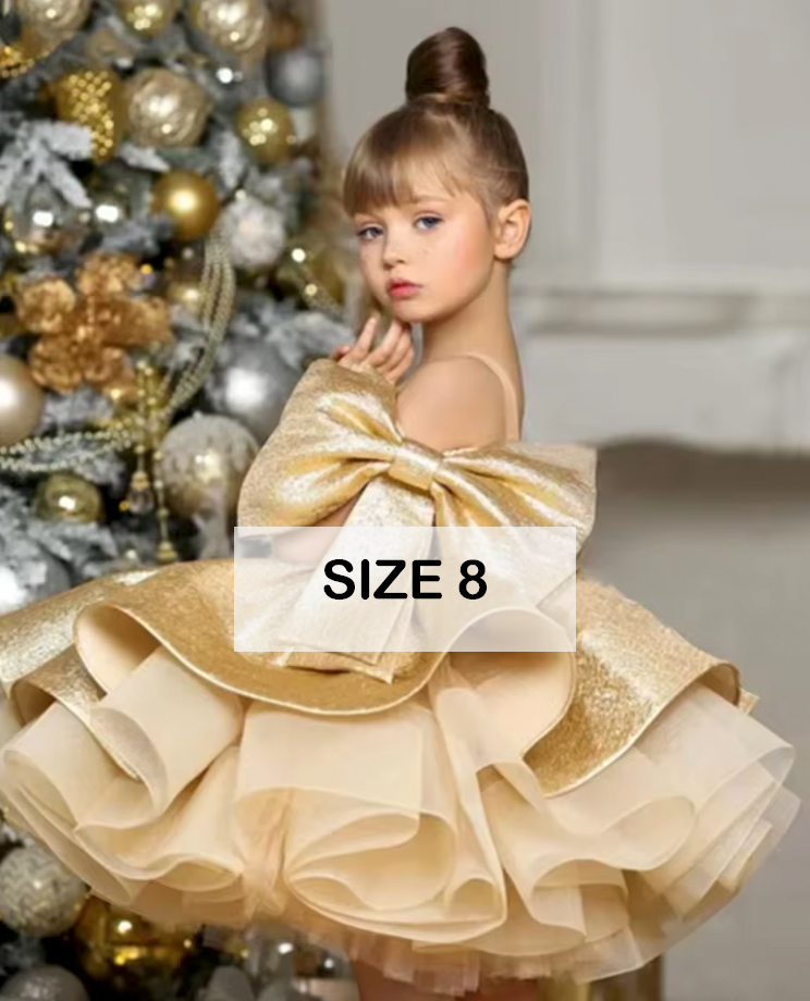 Girl wearing a gold glitter tiered ruffle ball gown dress with bow back, size 8, near Christmas tree.