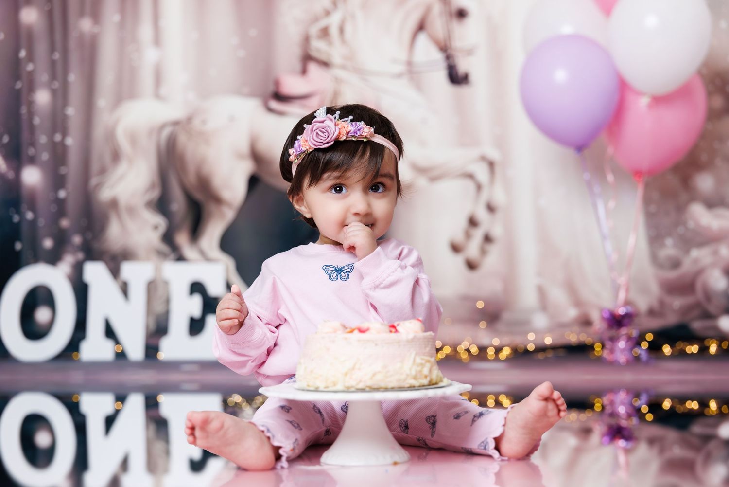 A baby girl tastes her first birthday cake surrounded by ONE signs and pink balloons.
