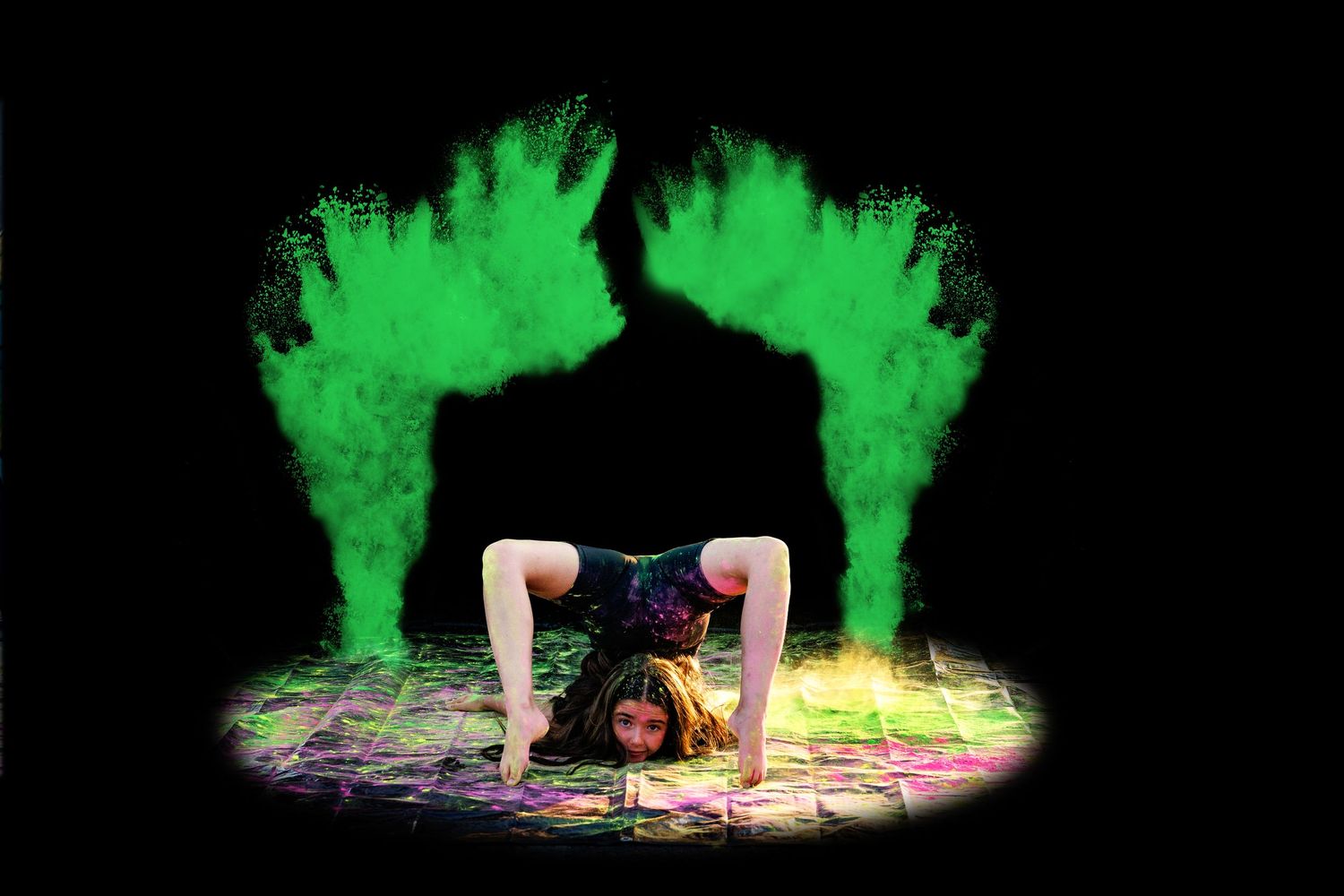 Flexible dancer performs backbend with twin green powder clouds rising on each side in darkness.