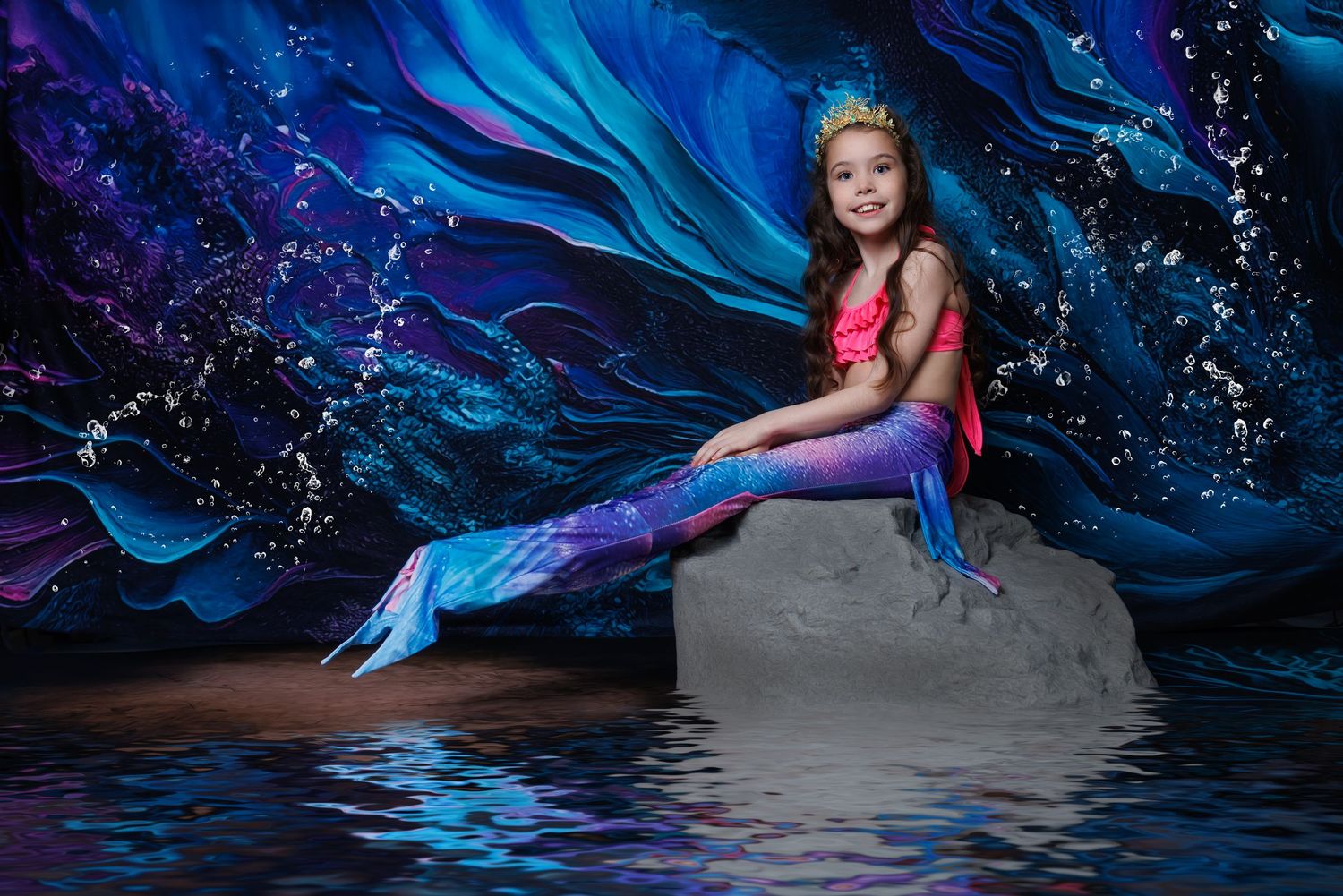 A young girl in a colorful mermaid tail and crown poses on a rock before a vivid blue ocean backdrop.