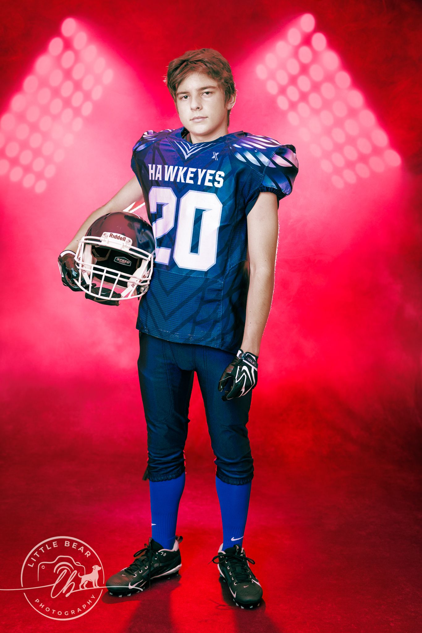 Youth Hawkeyes football player number 20 holding helmet under dramatic red stadium lights with smoky background.