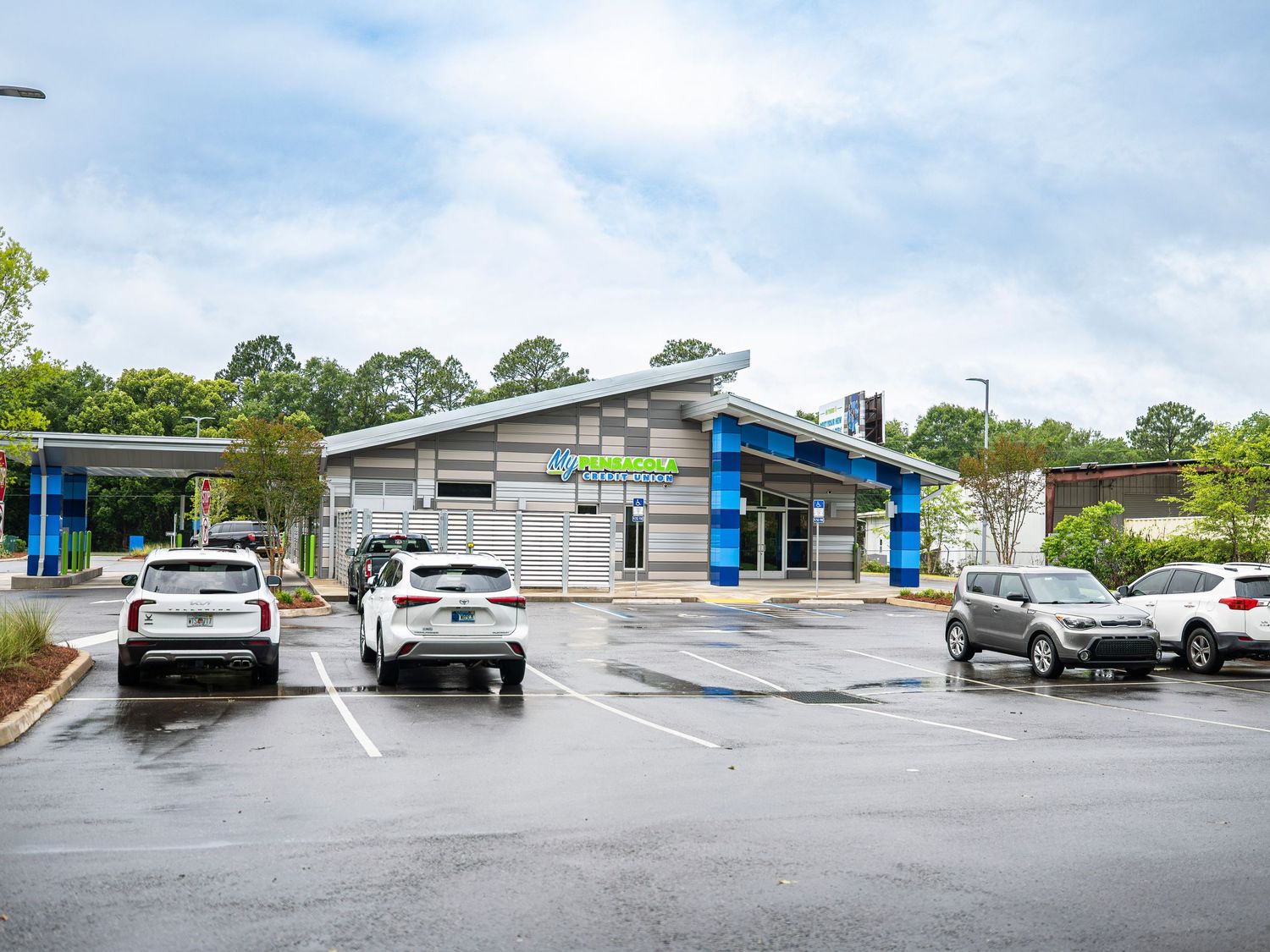 Modern credit union branch building with blue accents in a wet parking lot on a cloudy day.