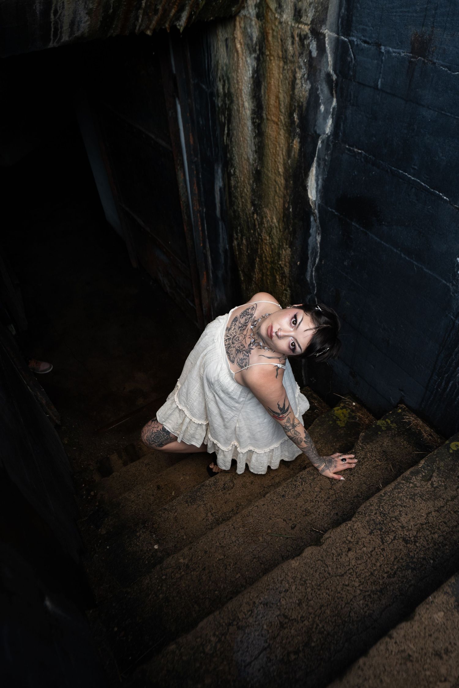Tattooed woman in white dress crawling up dark stone stairs in dramatic overhead perspective shot.