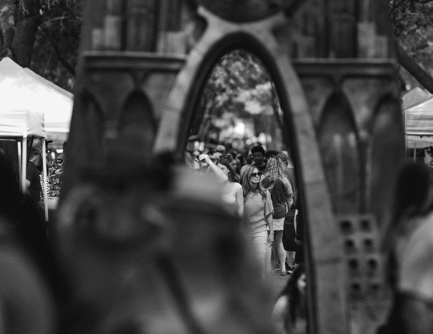 Black and white photo of crowd viewed through ornate gothic arch at outdoor market event.