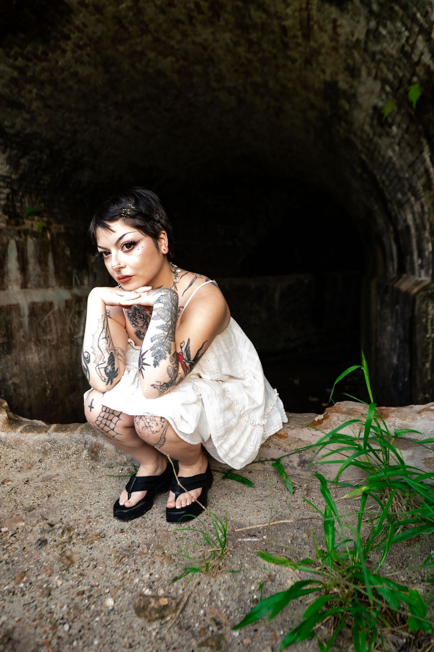 Tattooed woman in white dress crouching near dark tunnel entrance in outdoor urban setting.