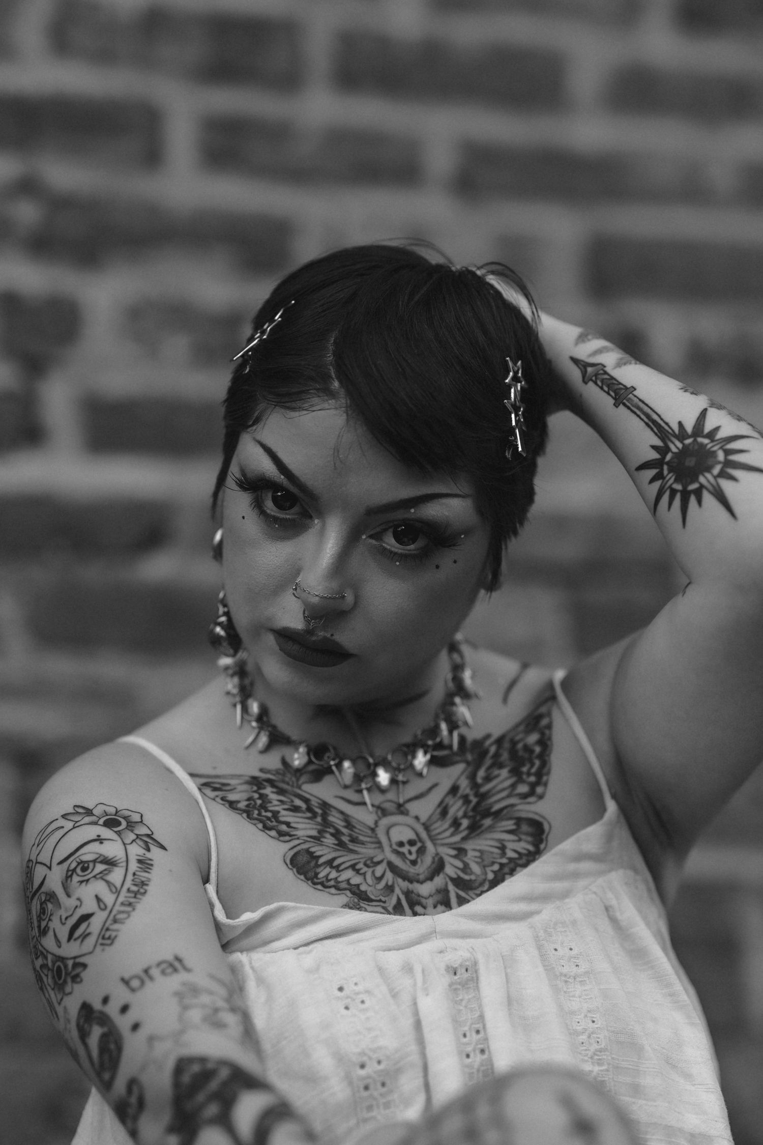 Close-up black and white portrait of tattooed woman with bold makeup and spiked necklace by brick wall.