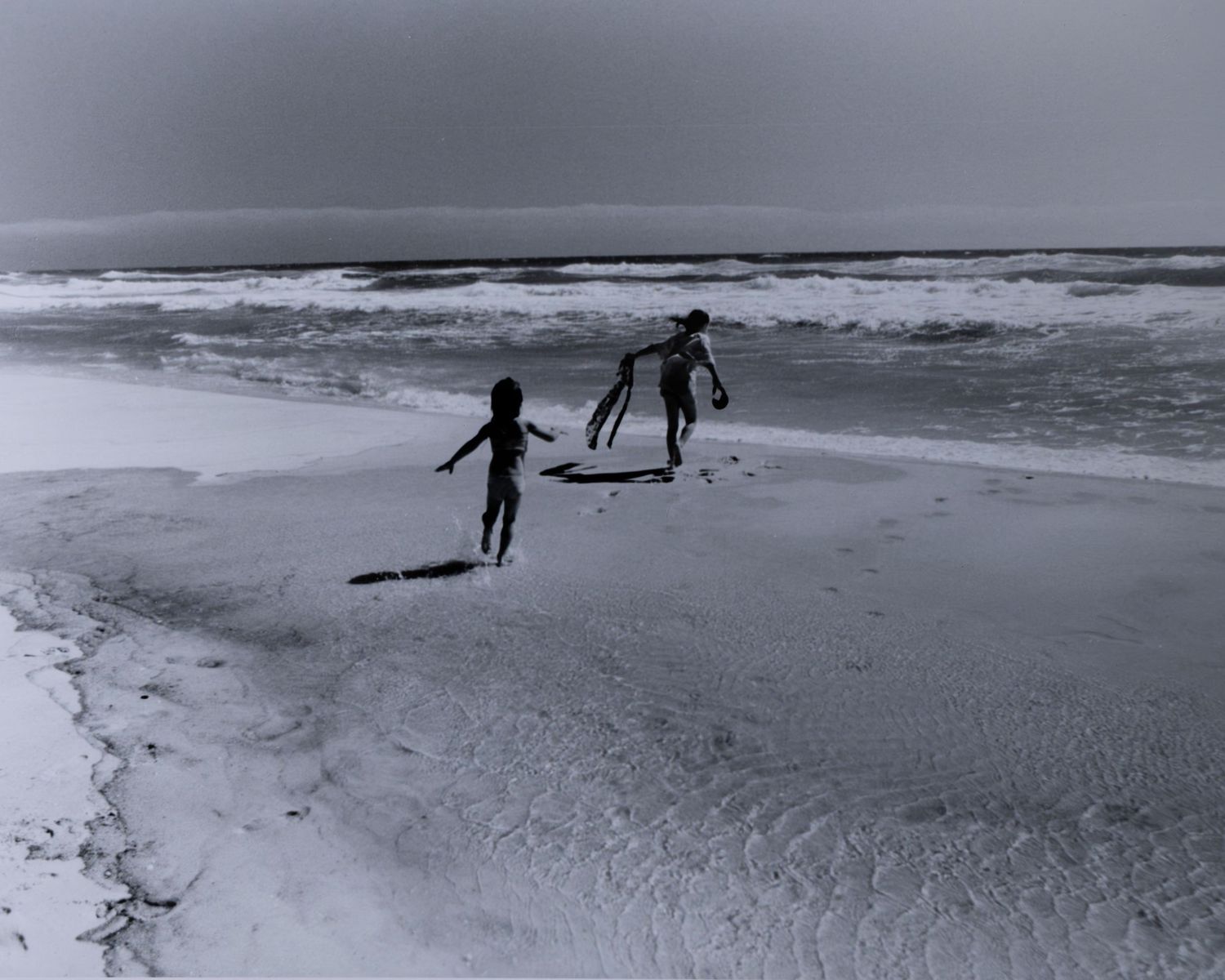 Two children running along a vast beach shoreline with ocean waves in a black and white photo.