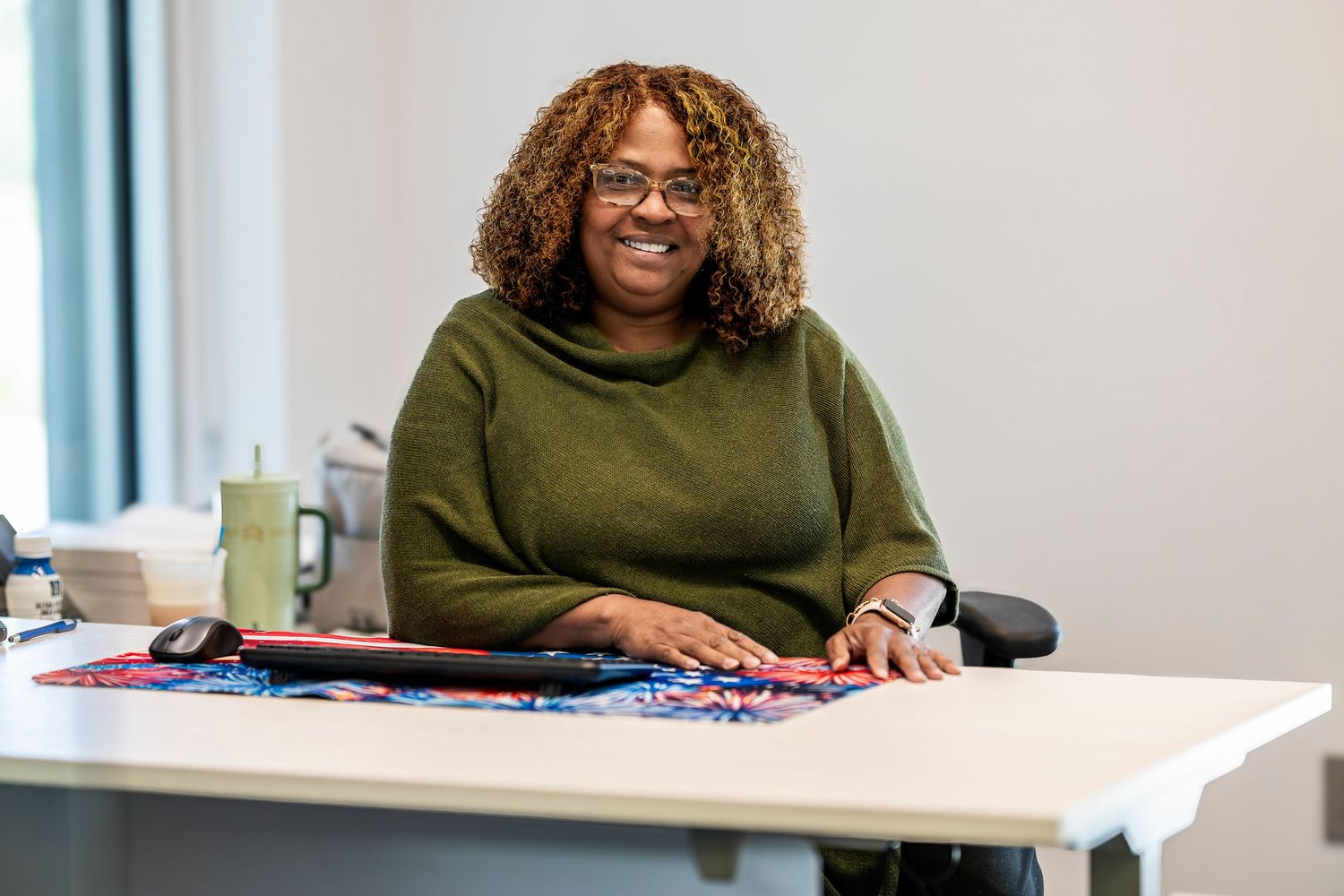 Smiling woman with curly hair and glasses sitting at a desk in a bright office.