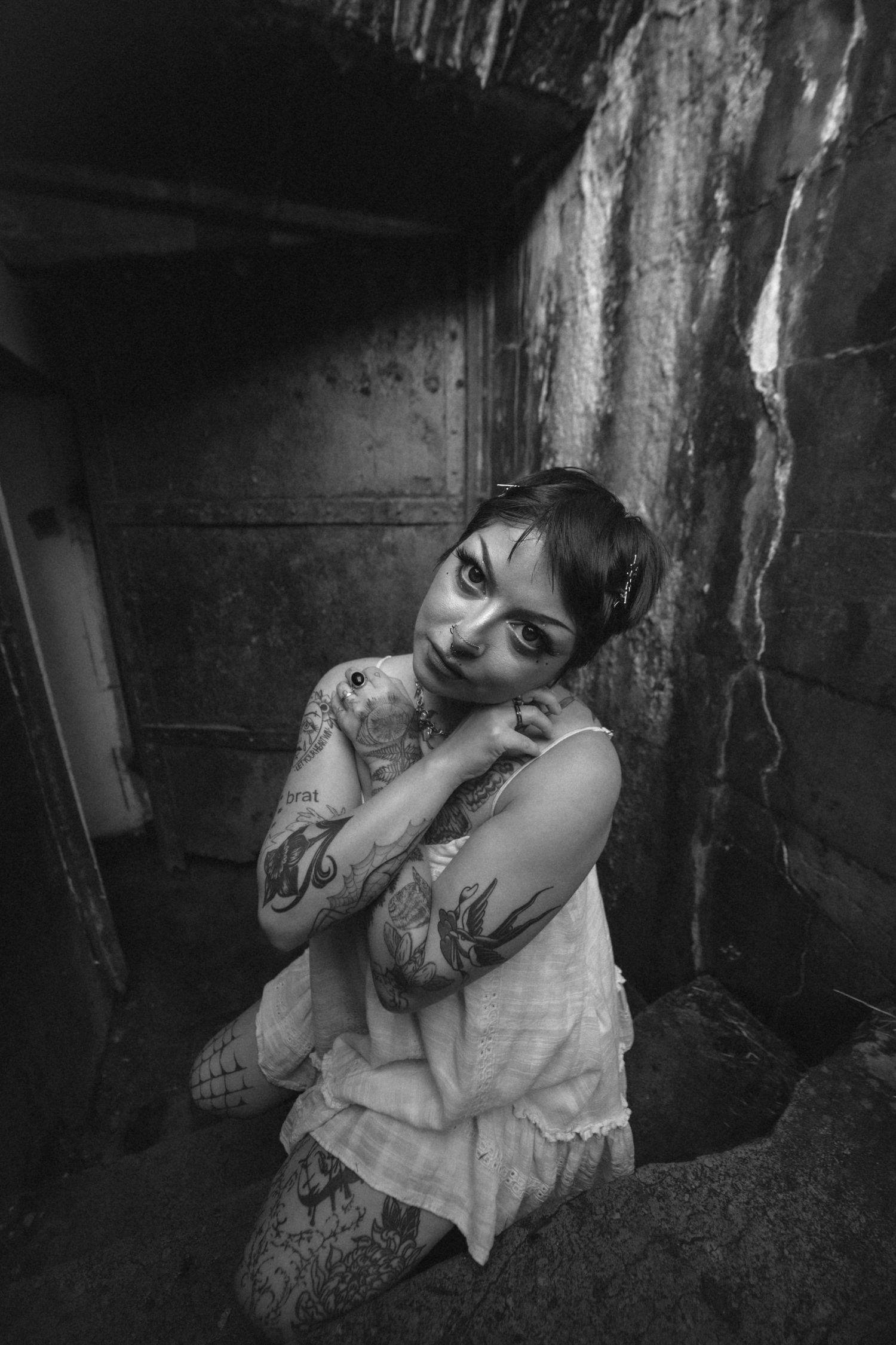 Tattooed woman in white dress crouching in dark abandoned building, black and white portrait photography.