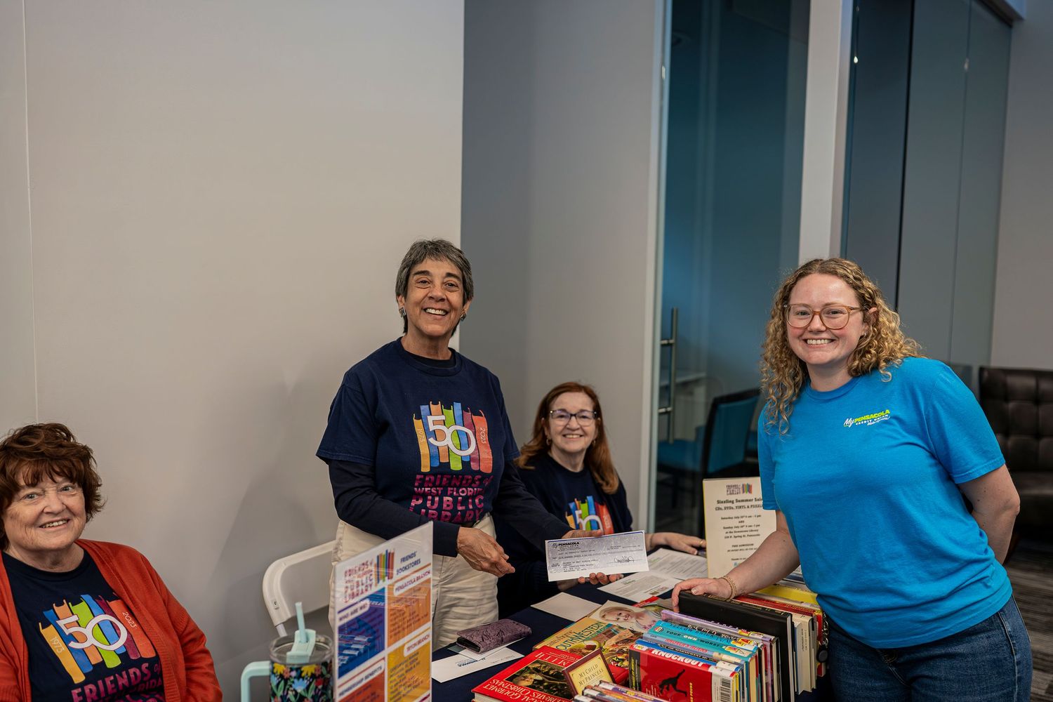 Four smiling volunteers stand behind a table filled with books at an indoor community literacy event.