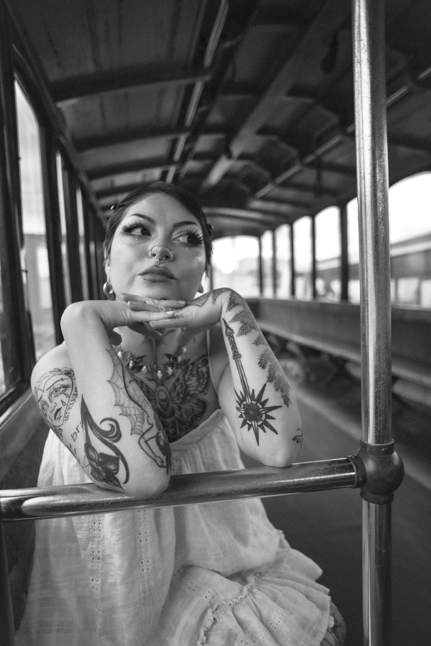Heavily tattooed woman in white dress leaning on metal rail inside vintage train car, black and white.