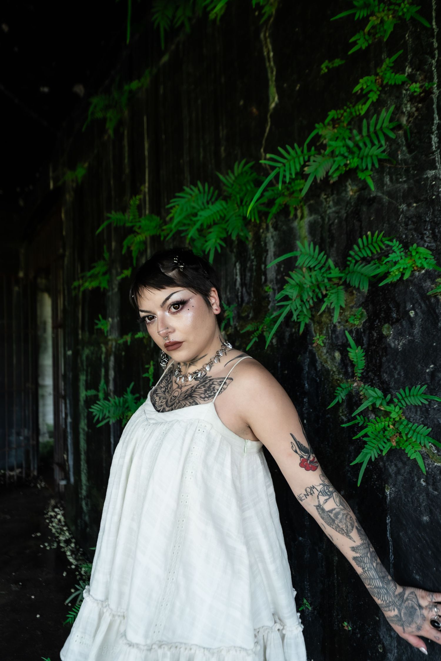 Tattooed woman in white sundress posing against dark wall with green ferns in moody portrait.