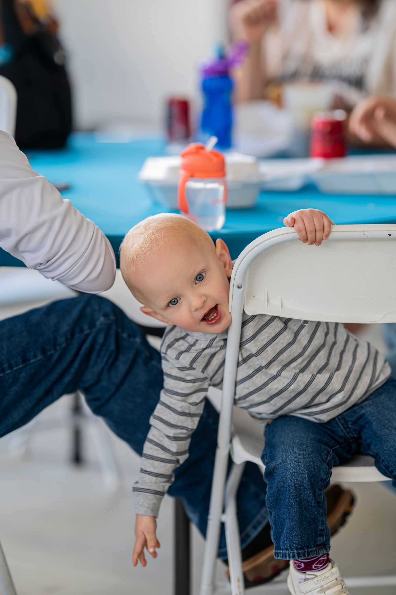 Happy baby in striped shirt leaning on a white chair at a family dining table.