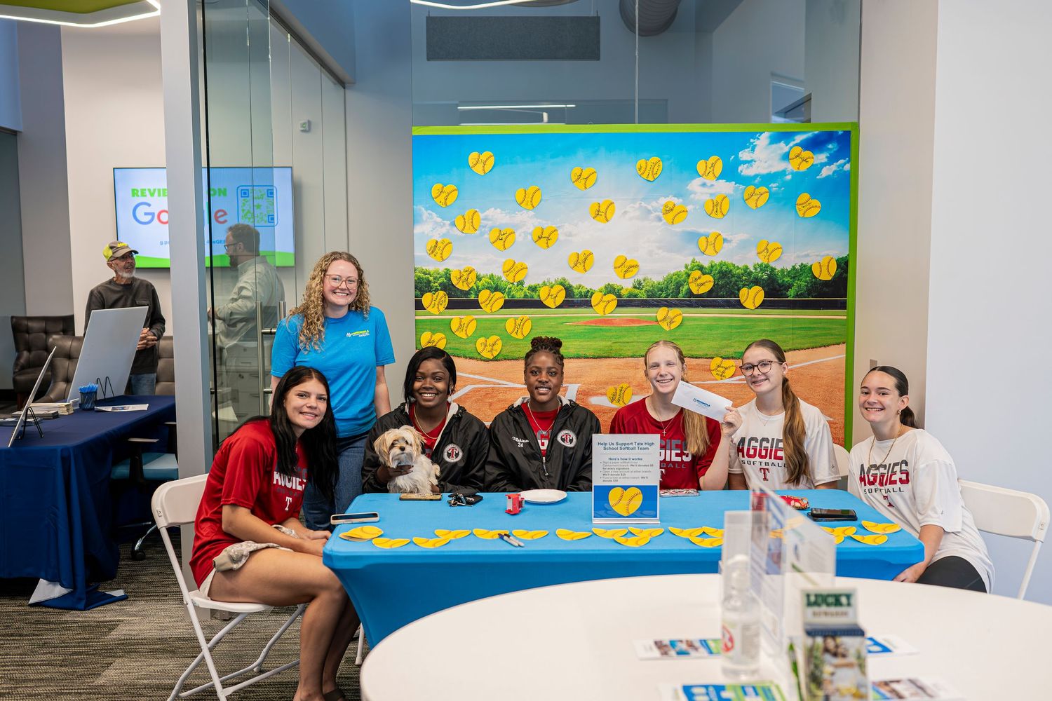 Group of smiling students pose at a decorated table with a small dog at an indoor school event.
