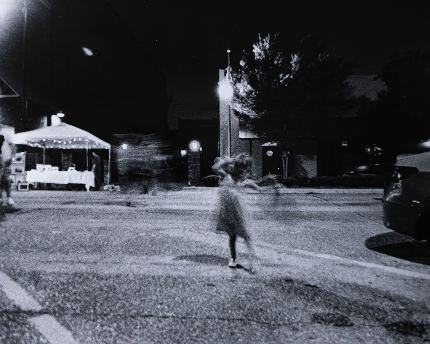 Blurry figure crossing a street at night near a lit vendor tent in a black and white long exposure photo.