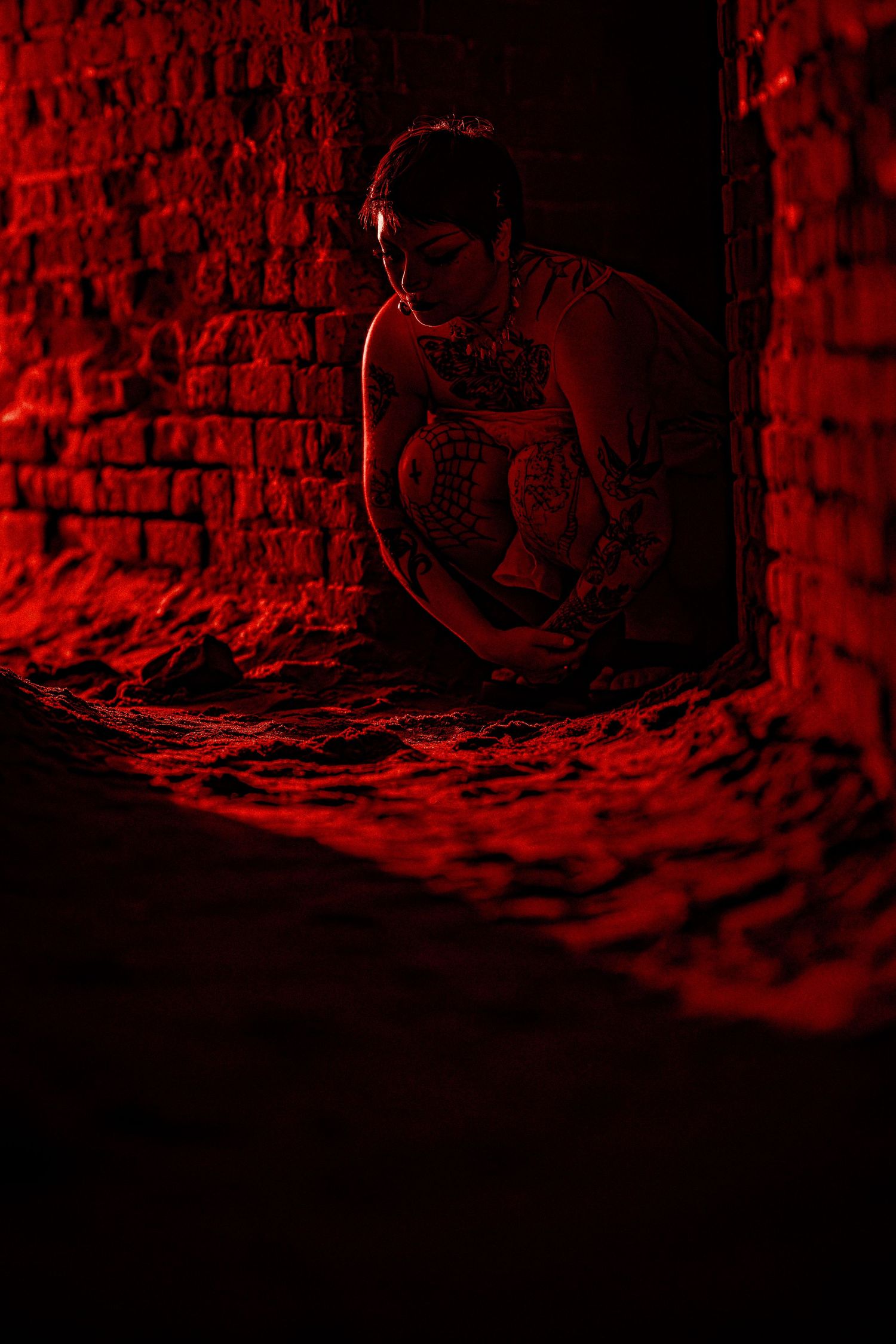 Tattooed woman sitting in brick doorway bathed in dramatic red light in dark abandoned space.