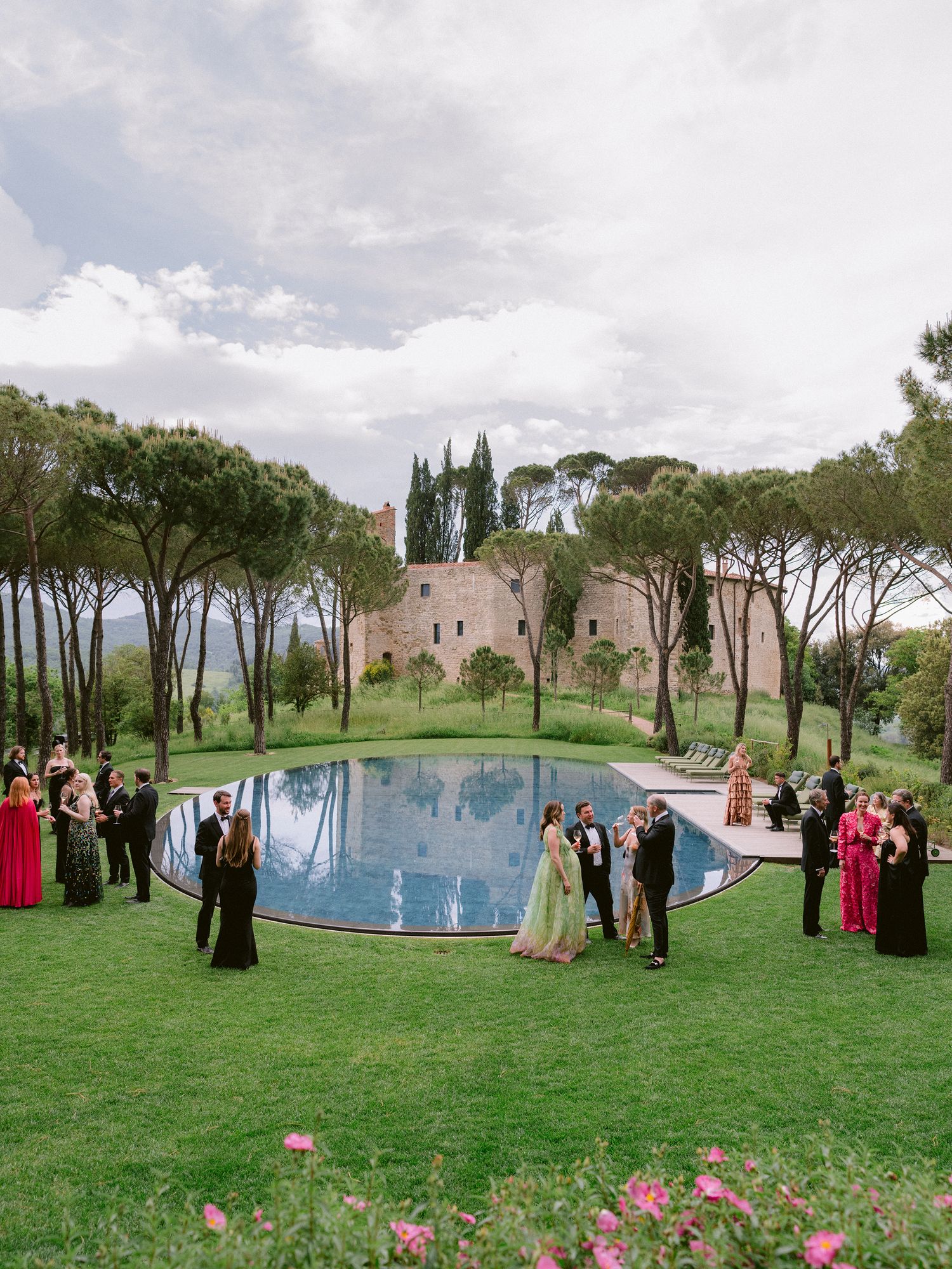 Wedding party gathers around an elegant outdoor pool surrounded by cypress trees and manicured gardens in Italy.
