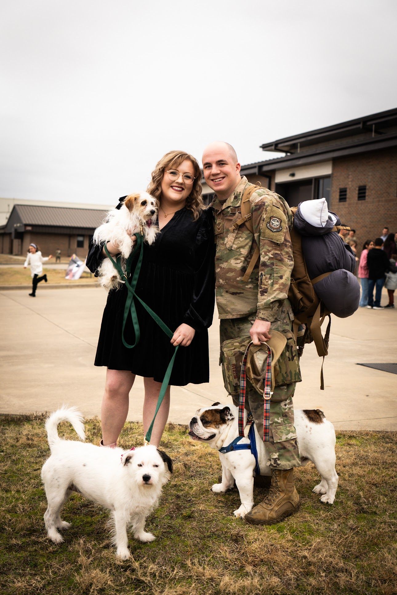 A smiling couple with three Jack Russell Terrier dogs at a military homecoming ceremony outdoors.