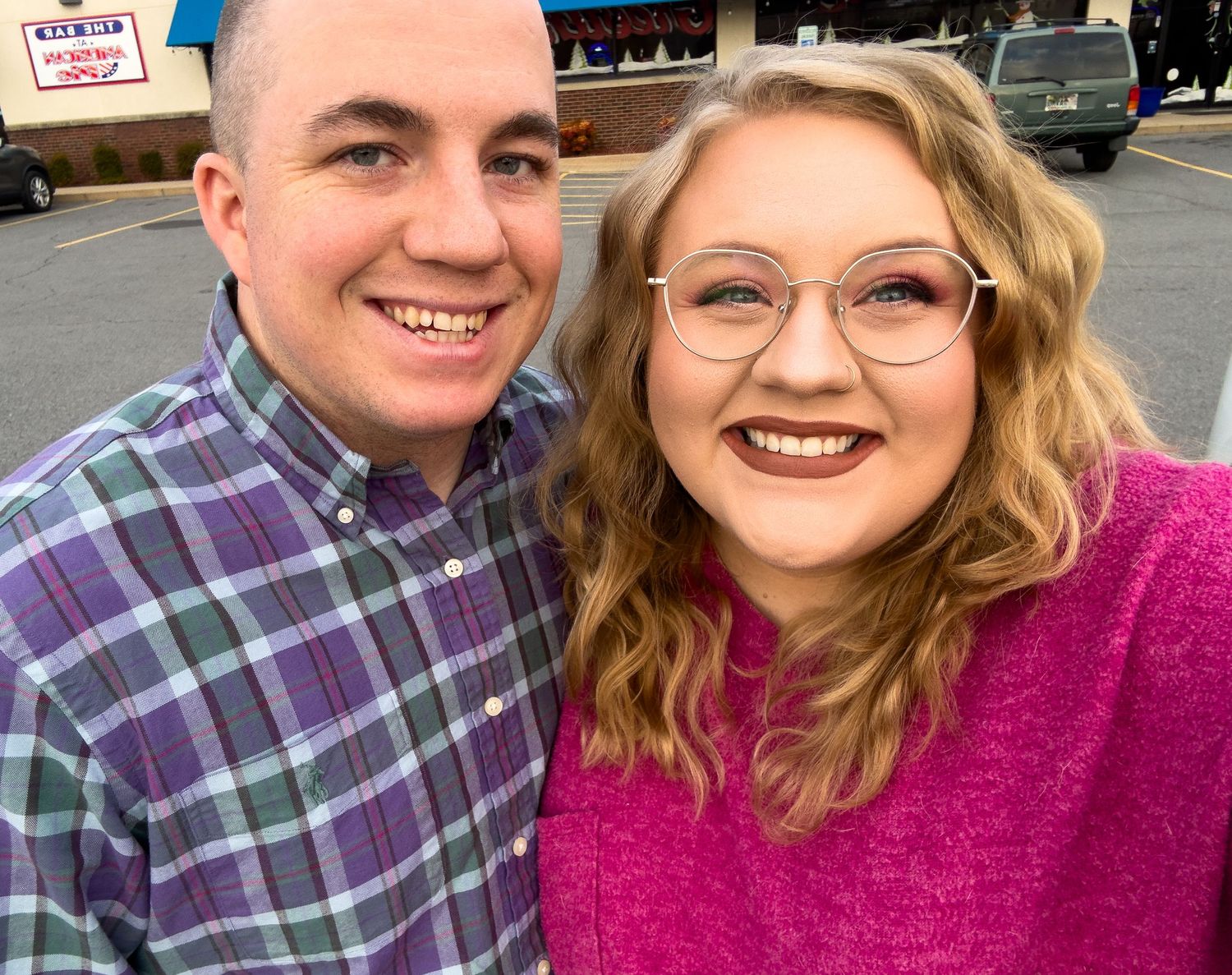 A smiling young couple taking a selfie outdoors, woman in pink sweater and glasses, man in plaid shirt.