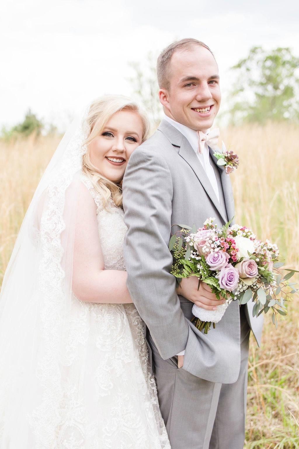 A happy bride and groom posing in a golden field, bride holding a pink rose bouquet.