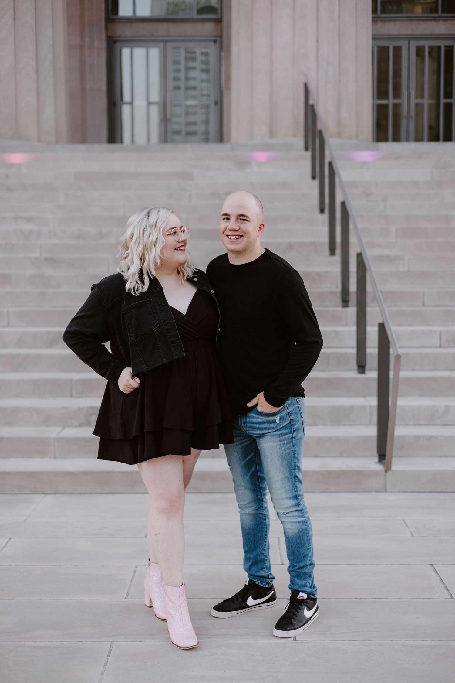 A smiling couple posing on stone steps, woman in black dress with pink boots, man in black sweater and jeans.