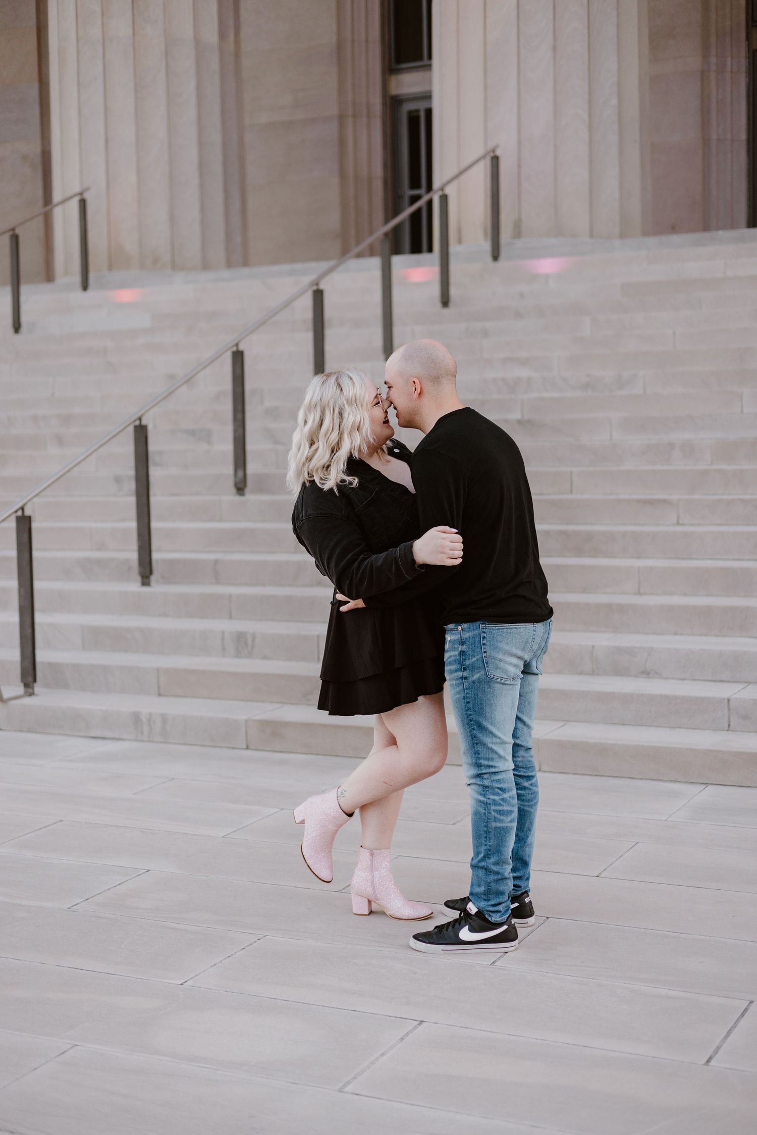 A couple sharing a romantic kiss on stone steps, woman in black dress with pink boots and man in jeans.