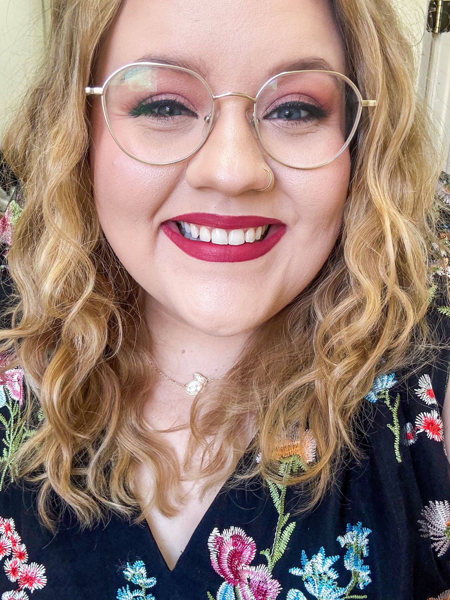 A smiling woman with curly blonde hair, round glasses, bold red lips, and colorful eye makeup.