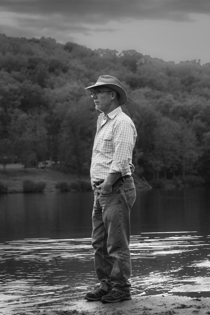 Black-and-white portrait of a man wearing a brimmed hat beside a lake, with wooded hills behind him.