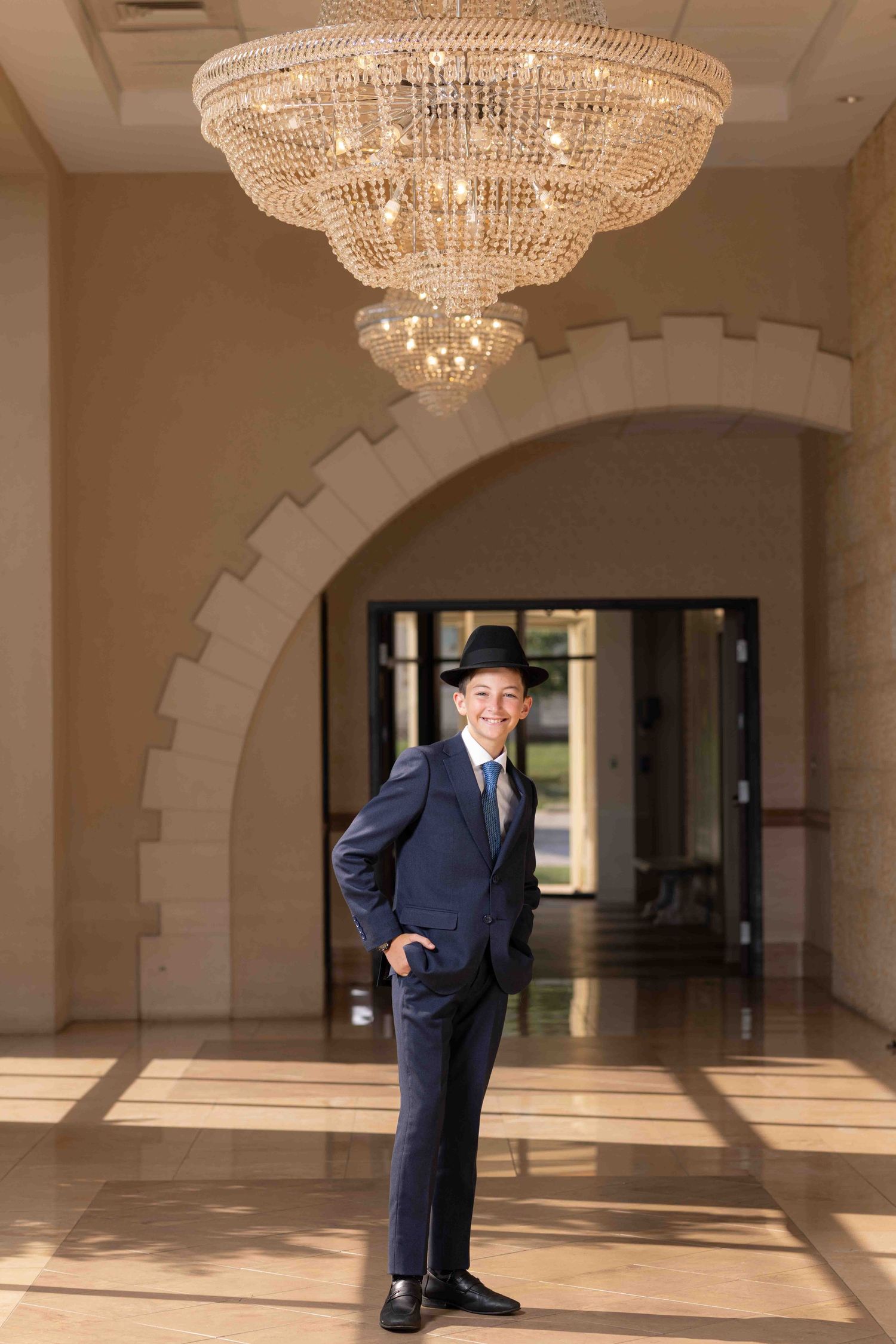 Young boy in navy suit and black fedora hat smiling under crystal chandelier in elegant venue hallway.