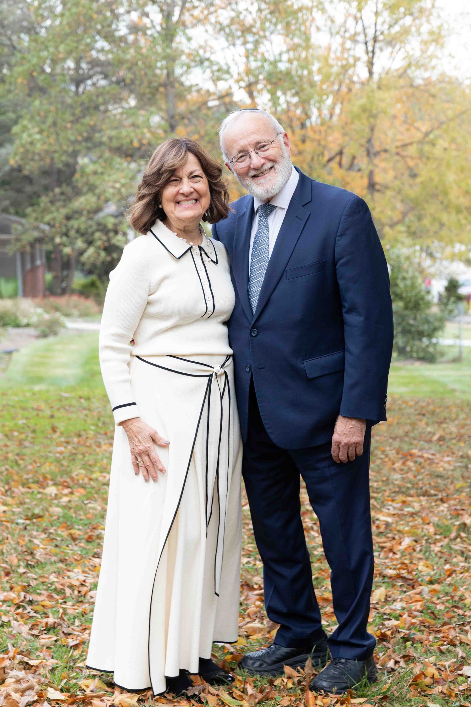 Smiling older couple in formal wear poses together outdoors among autumn leaves in a garden.