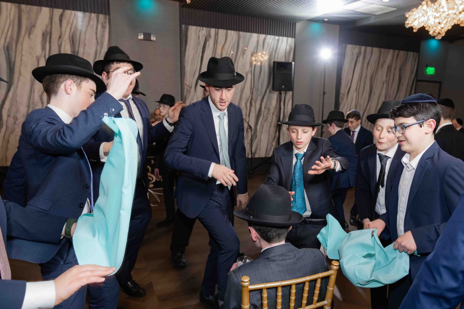 Group of young men in suits and hats dance energetically with teal ties at a Jewish wedding.