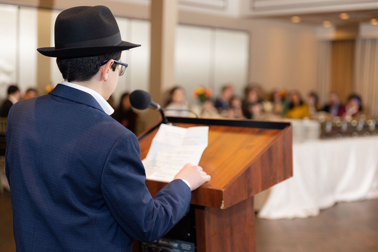 Rabbi in black fedora reads from podium at Bar Mitzvah reception with seated guests.