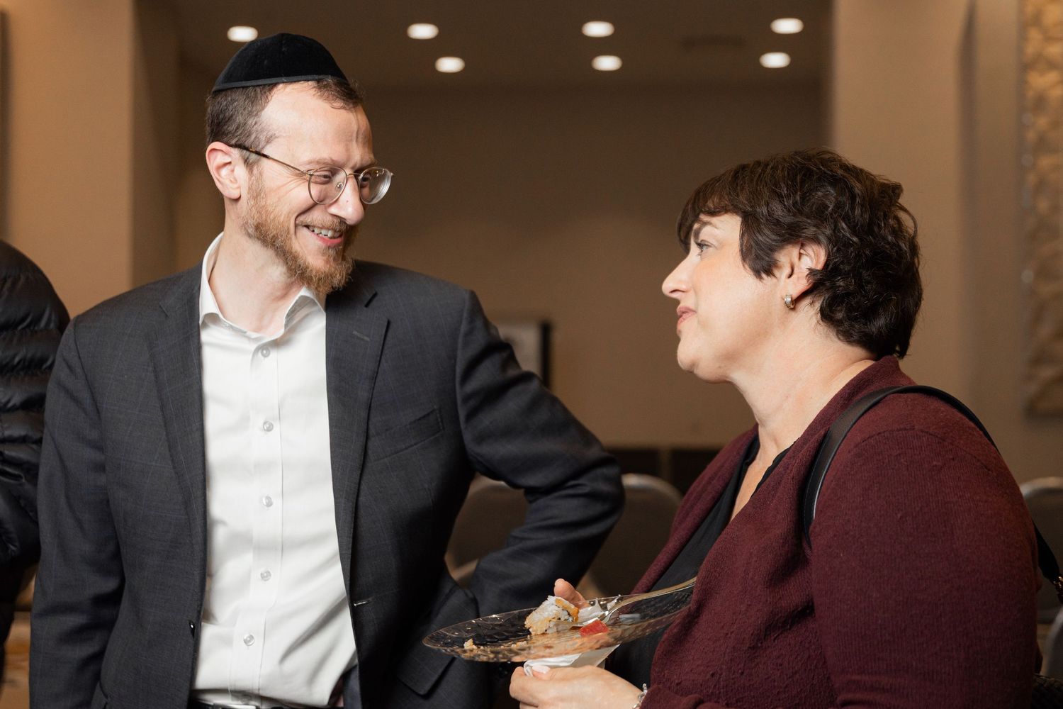 A man wearing a kippah smiles while conversing with a woman at a Jewish event.