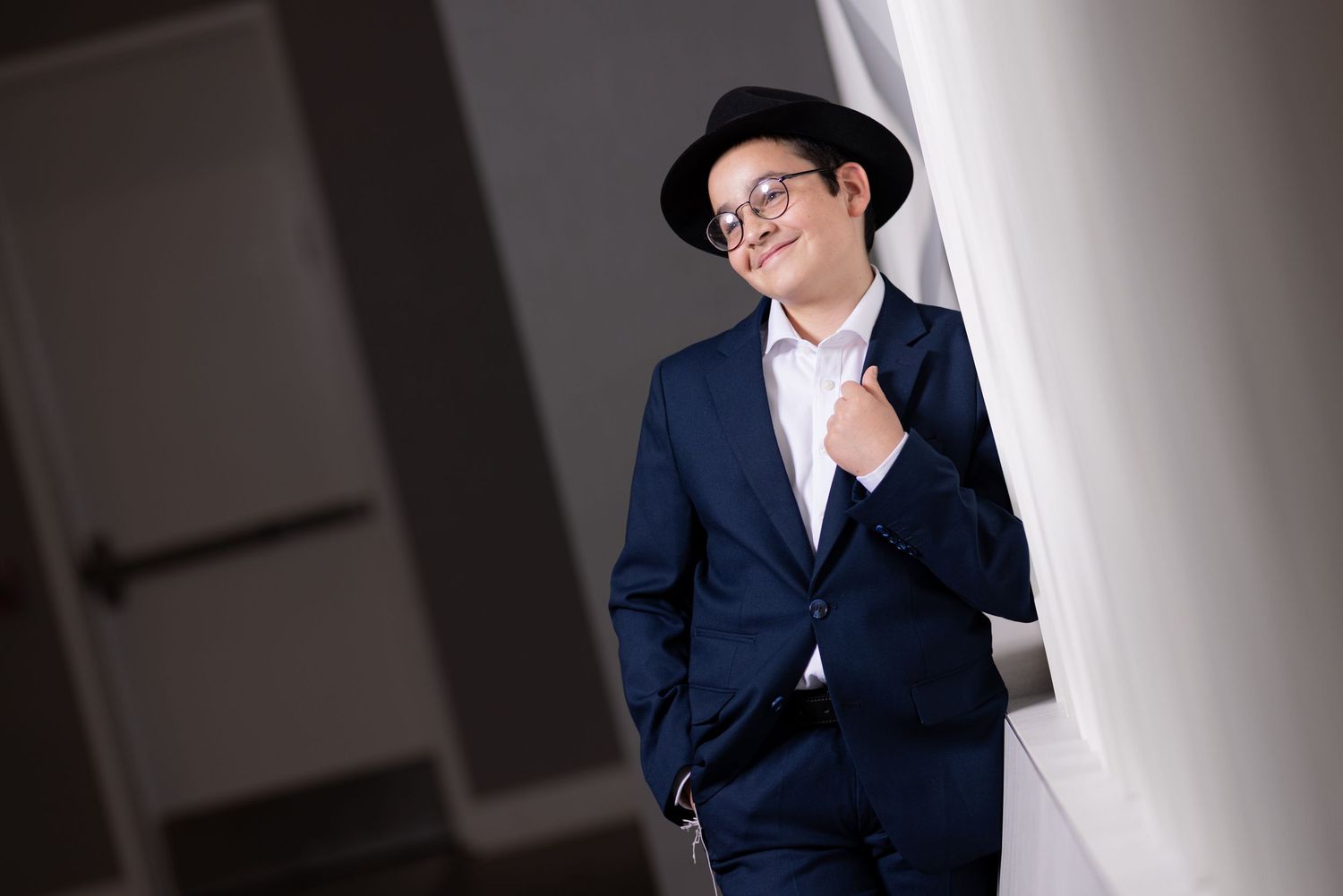 Young man in blue suit and black hat leaning against white curtain, smiling confidently indoors.