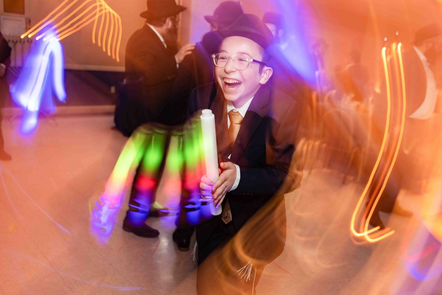 A joyful boy in a suit and hat dances with glowing light sticks at a lively Jewish celebration.