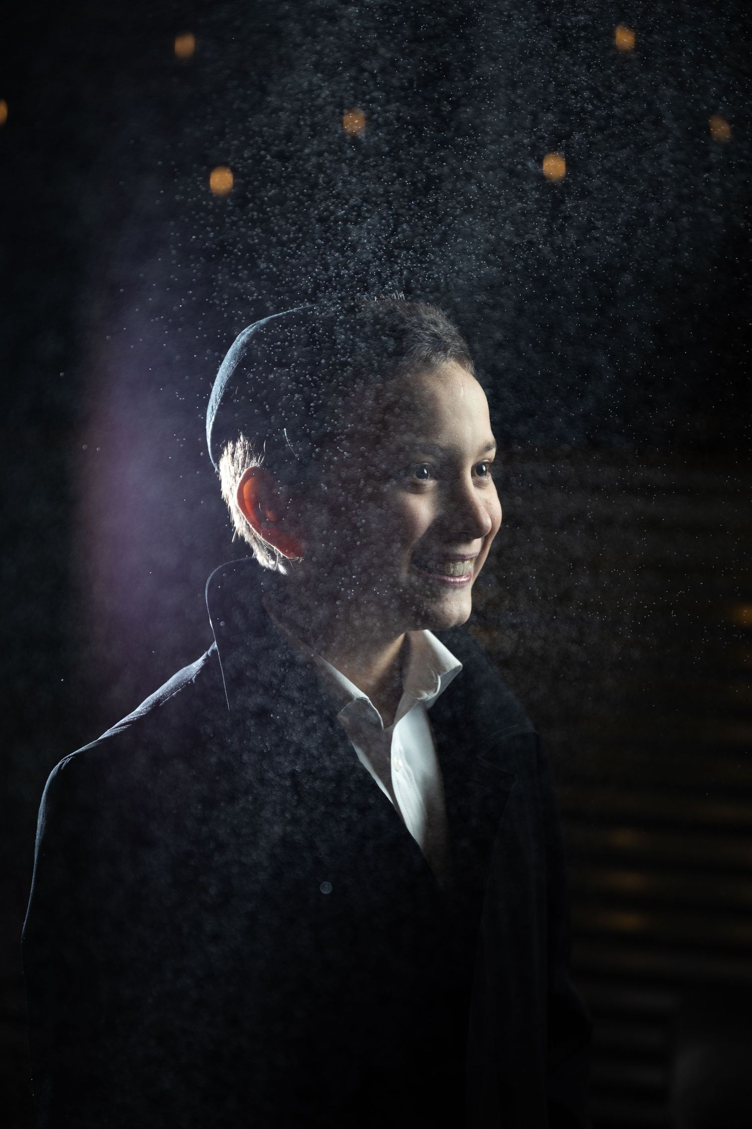 A smiling man in a dark suit is illuminated dramatically against a dark background with bokeh lights.