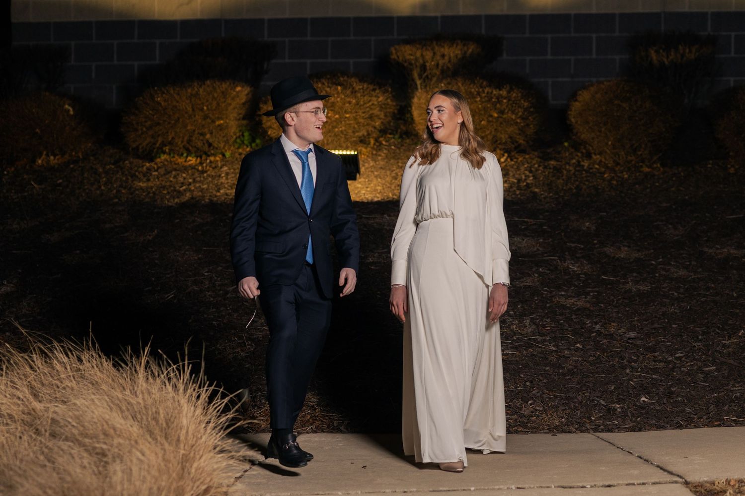 A man in a suit and hat walks alongside a woman in an elegant white dress outdoors at night.