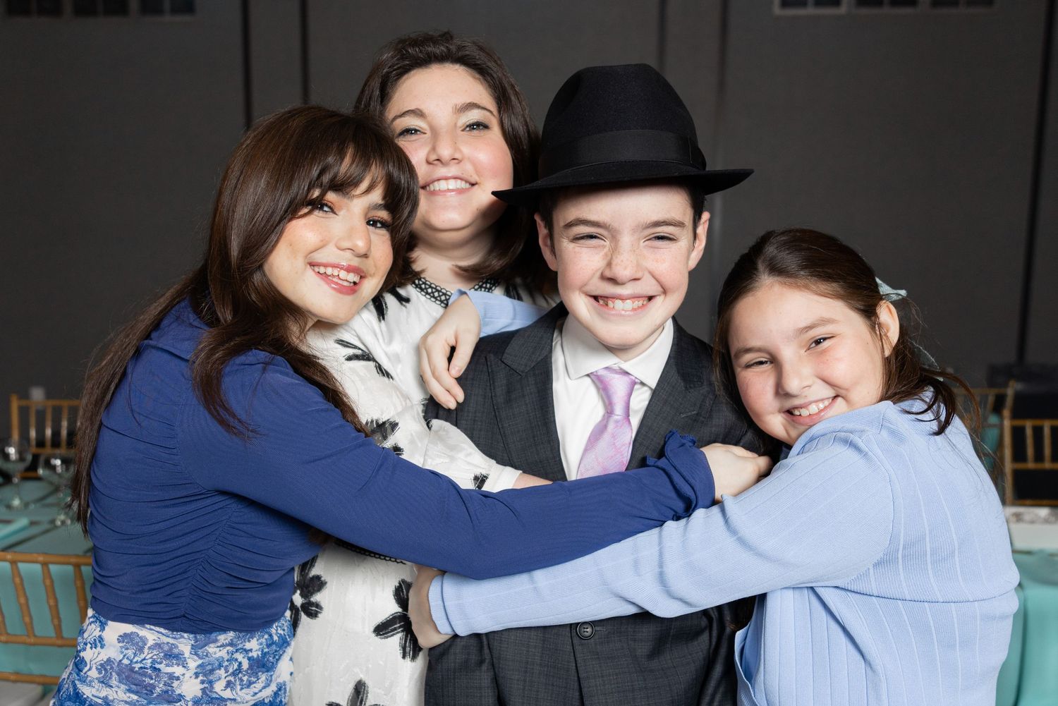 A boy in a black hat and suit smiles as three girls hug him at a Bar Mitzvah celebration.