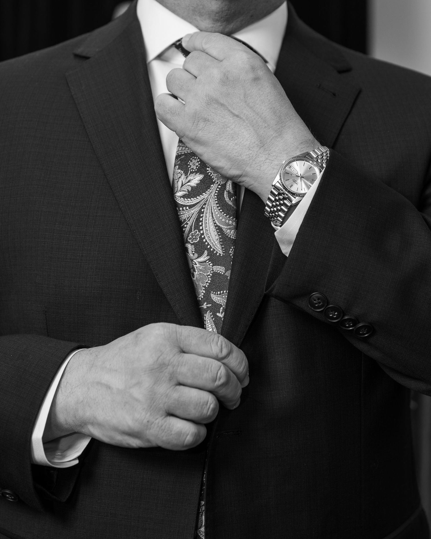 Black and white close-up of groom in suit adjusting paisley tie with luxury watch on wrist.