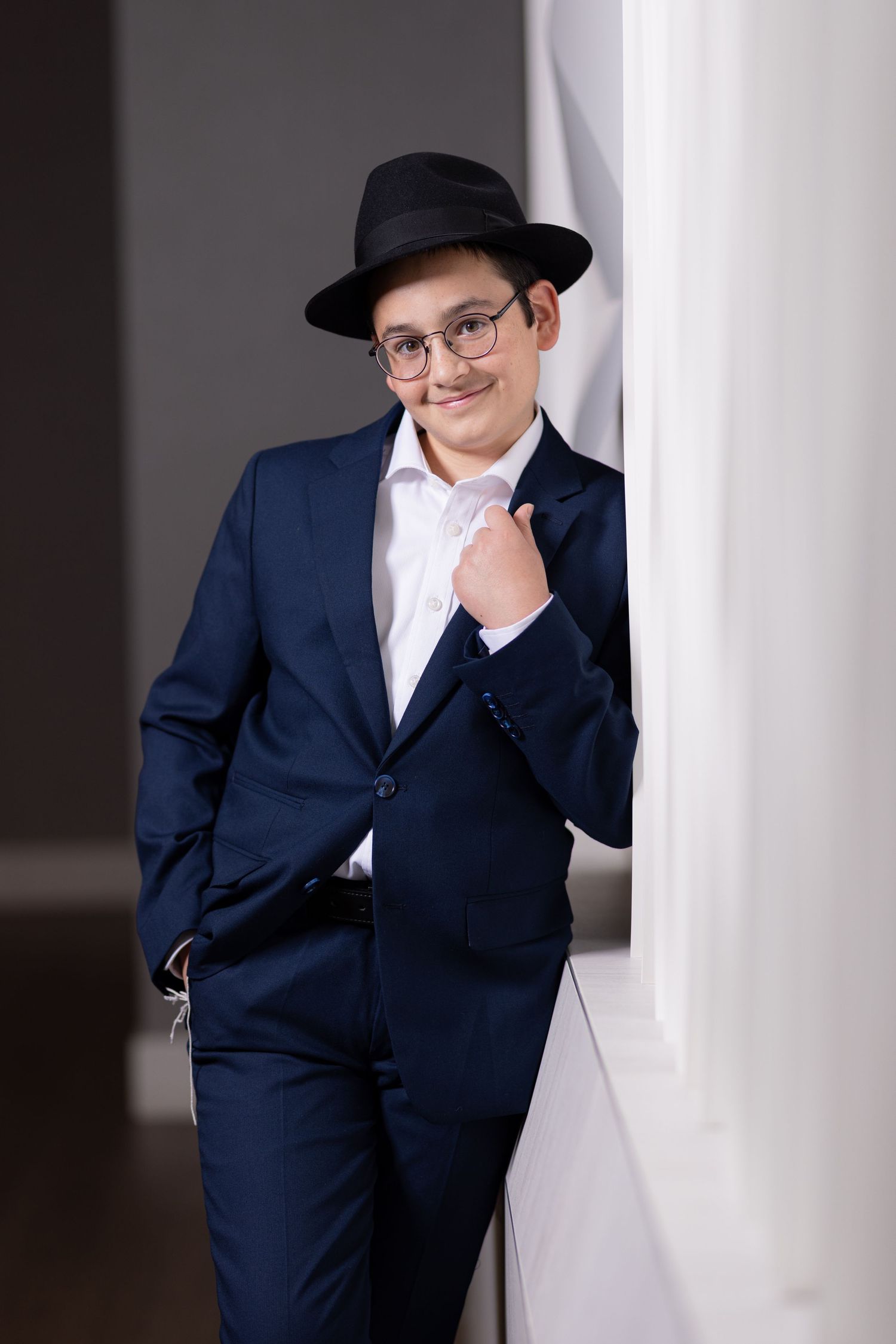 Young boy in navy suit, black fedora hat, and glasses poses confidently leaning against a white pillar.