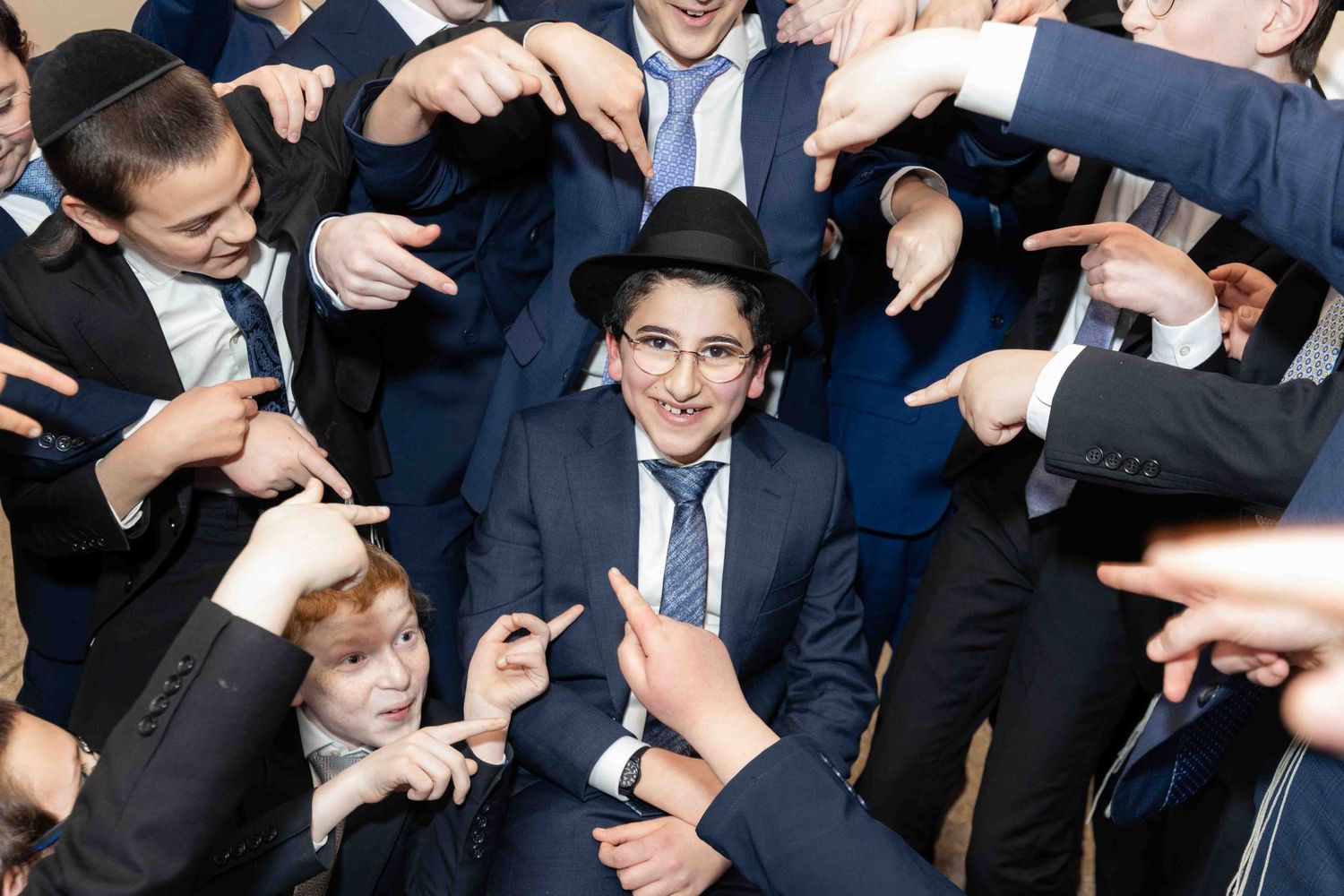 Smiling young man in suit surrounded by friends all pointing at him at a Jewish celebration.