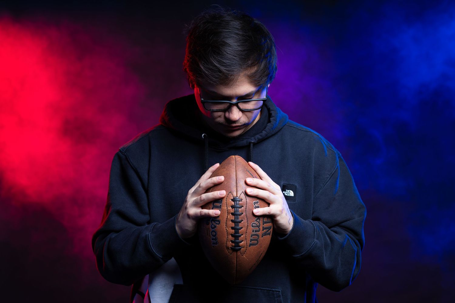 Teen boy in hoodie holds football against dramatic red and blue smoke background.
