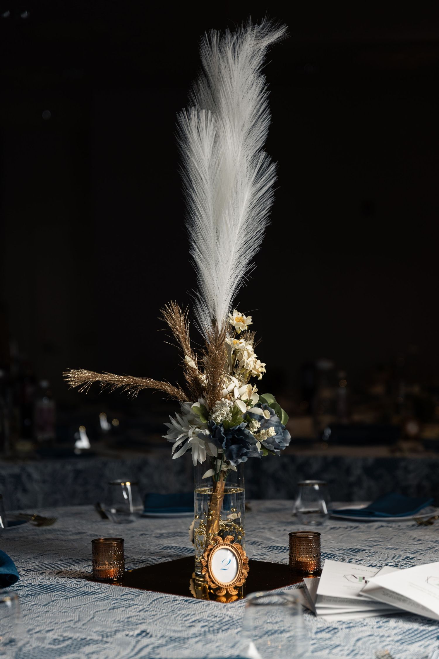 Elegant wedding centerpiece with white feather plume, dried flowers, and gold frame on lace tablecloth.