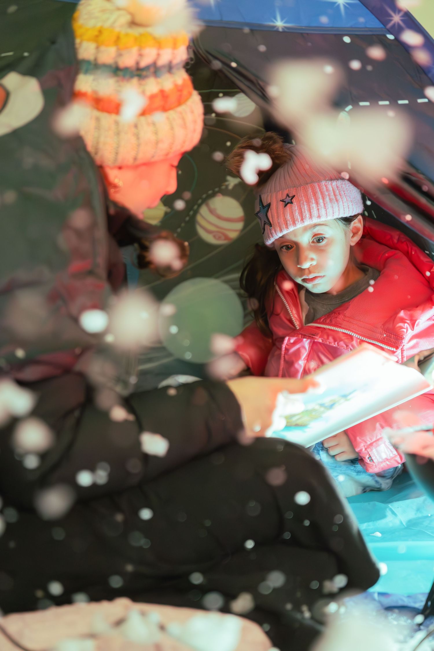 A young girl in a pink winter hat reads a book inside a car during a snowy night.