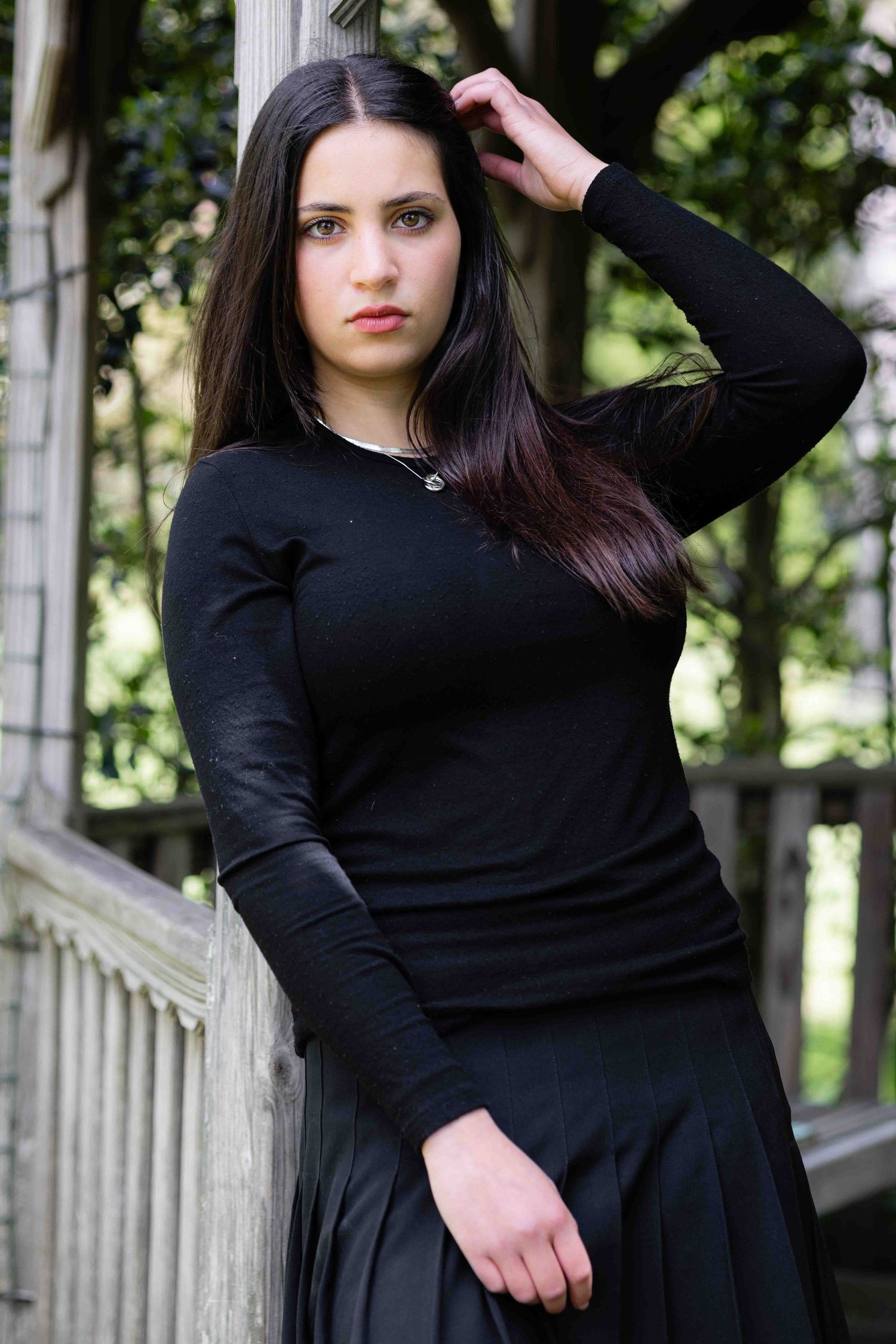 Young woman with long dark hair in black outfit leaning against gazebo pillar outdoors.