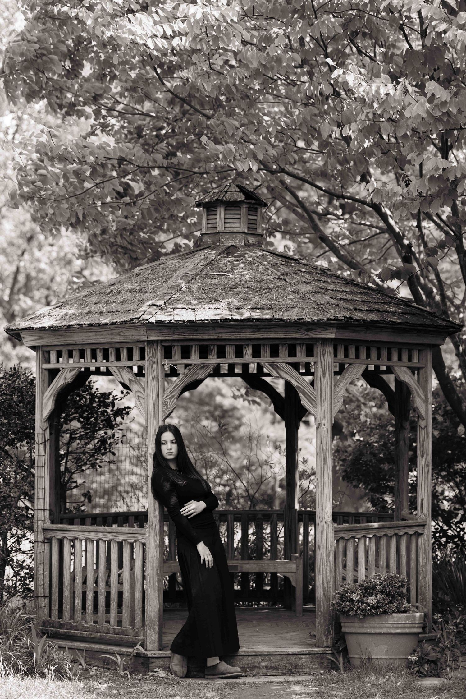 Black and white photo of young woman in black dress posing by rustic wooden gazebo.