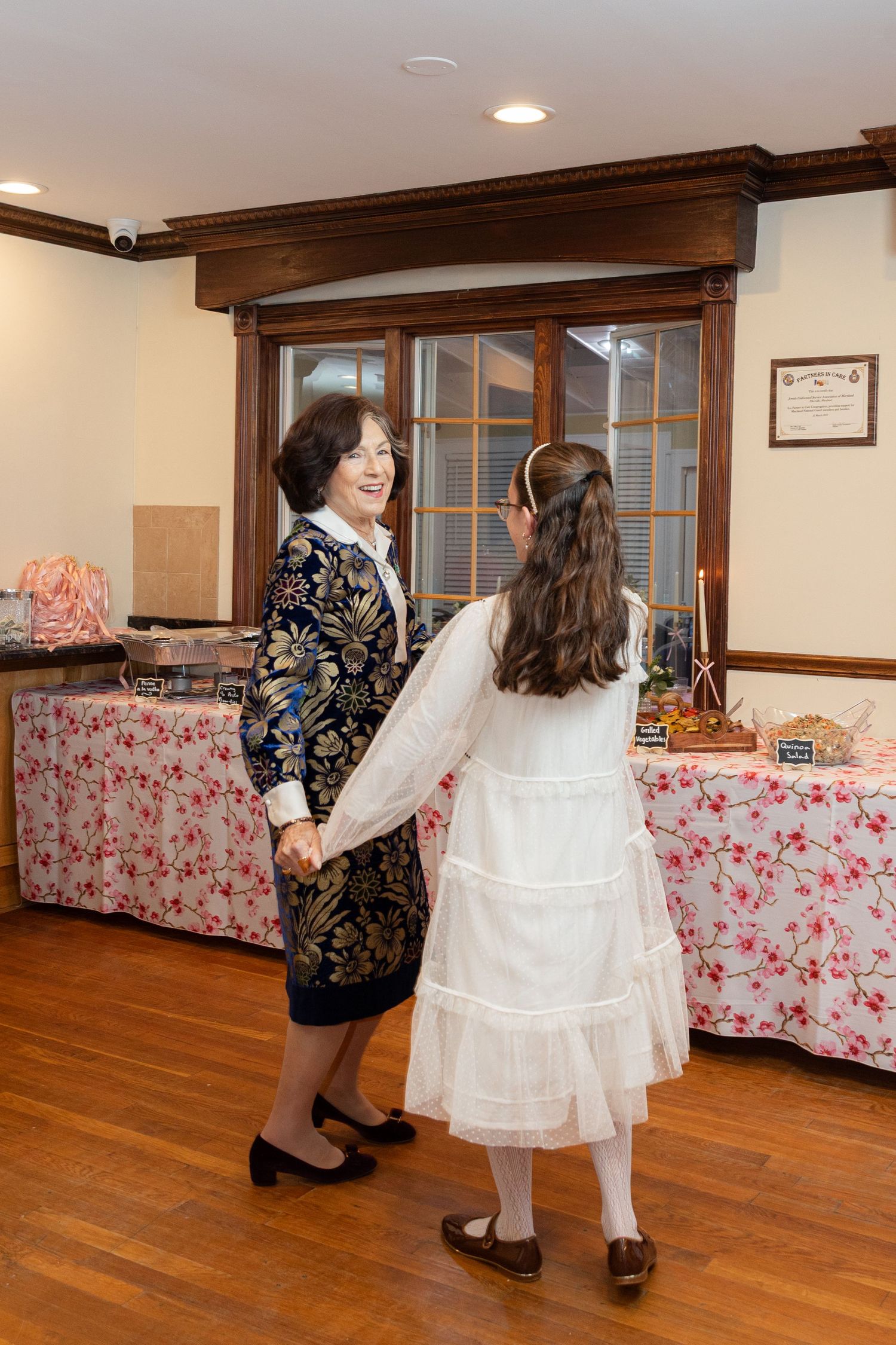 An elderly woman and a young girl in white dress hold hands and dance at a family celebration.