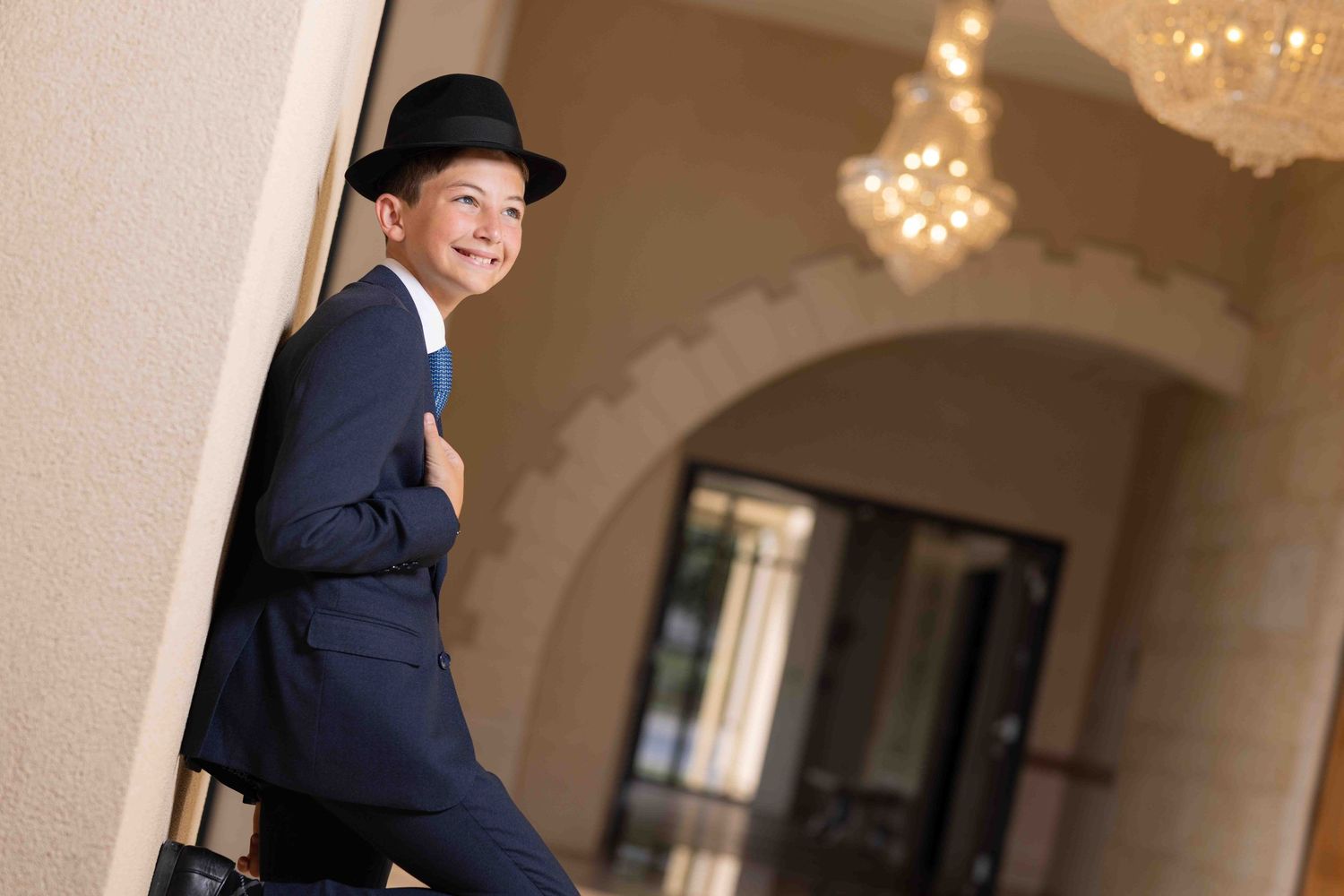Smiling boy in navy suit and black bowler hat leaning against wall in elegant venue with chandelier.